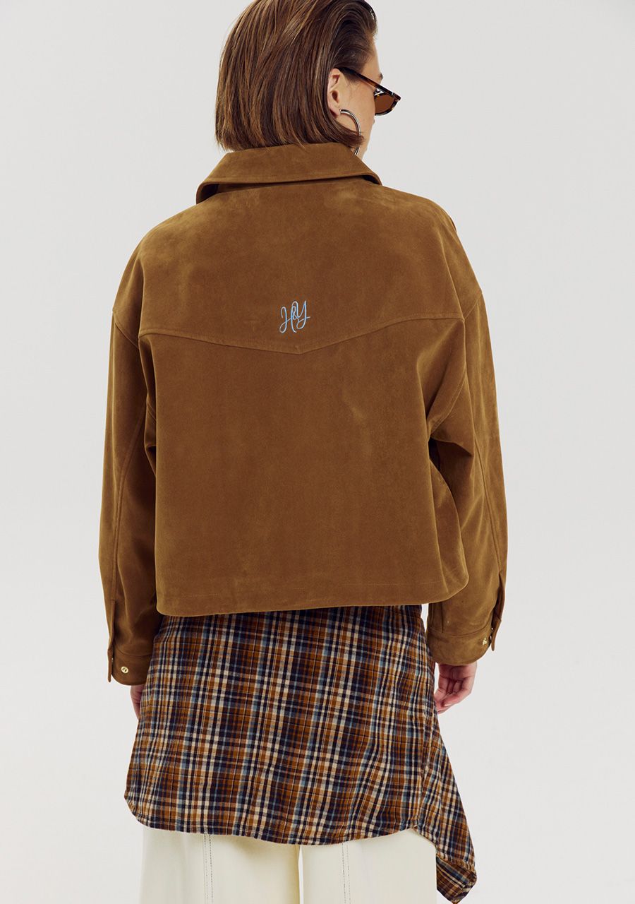 A model shown from the back wearing a cropped brown suede jacket with a small embroidered logo on the upper back, paired with a brown and beige plaid shirt and white pants, against a light gray studio background.