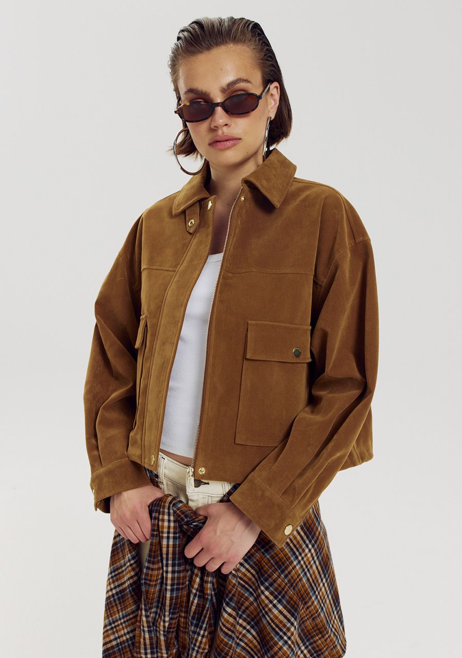A model wearing a brown suede utility jacket over a white top and a plaid skirt, styled with dark sunglasses, against a light gray studio background.