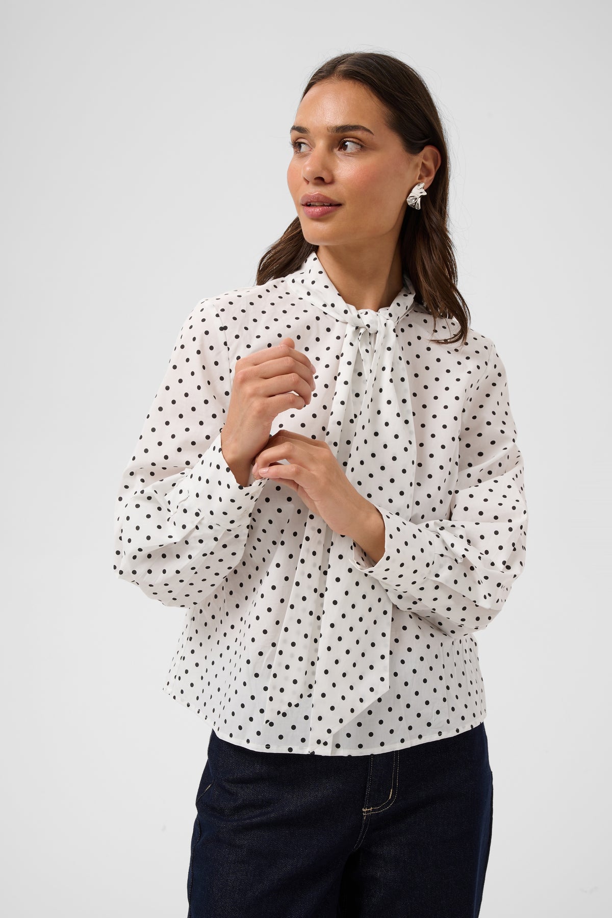 A woman wearing a white long-sleeve blouse with a black polka dot pattern and a tie-neck detail, paired with dark blue jeans, against a light gray studio background.