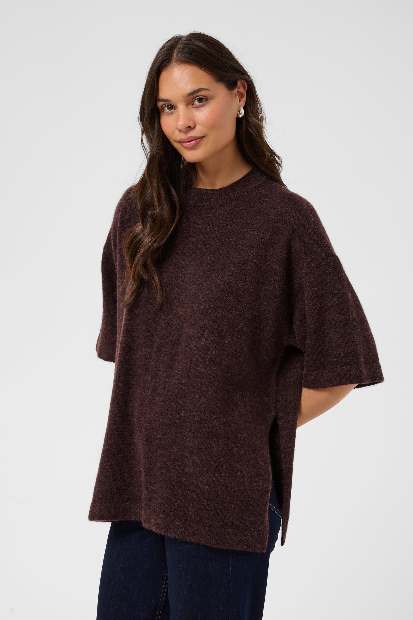 A woman modeling an oversized short-sleeve knit top in a dark plum-brown color, paired with dark blue jeans, against a light gray studio background.