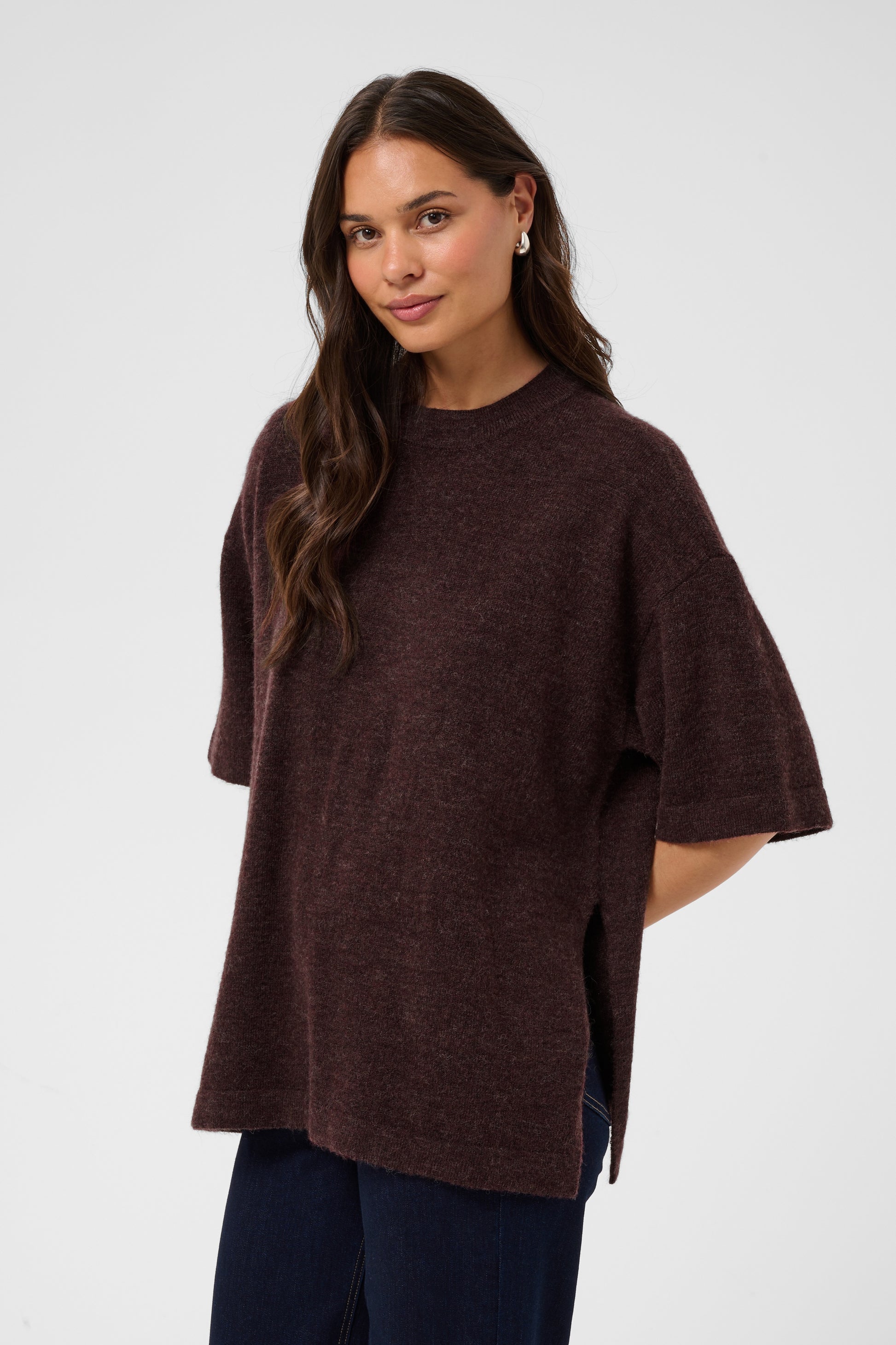 A woman modeling an oversized short-sleeve knit top in a dark plum-brown color, paired with dark blue jeans, against a light gray studio background.