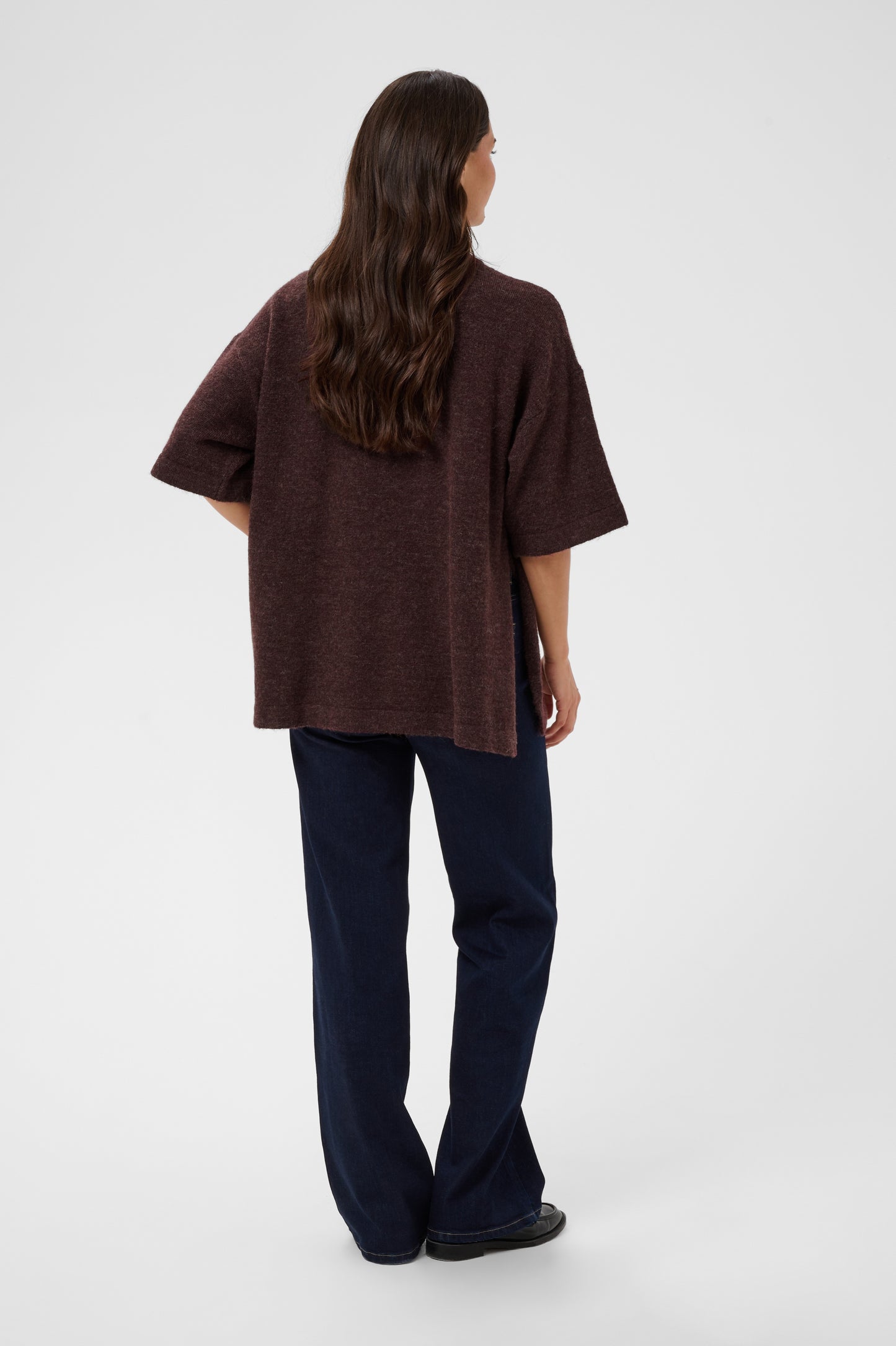 A woman viewed from the back wearing an oversized dark brown short-sleeve knit top and dark navy pants, standing against a light gray studio background.