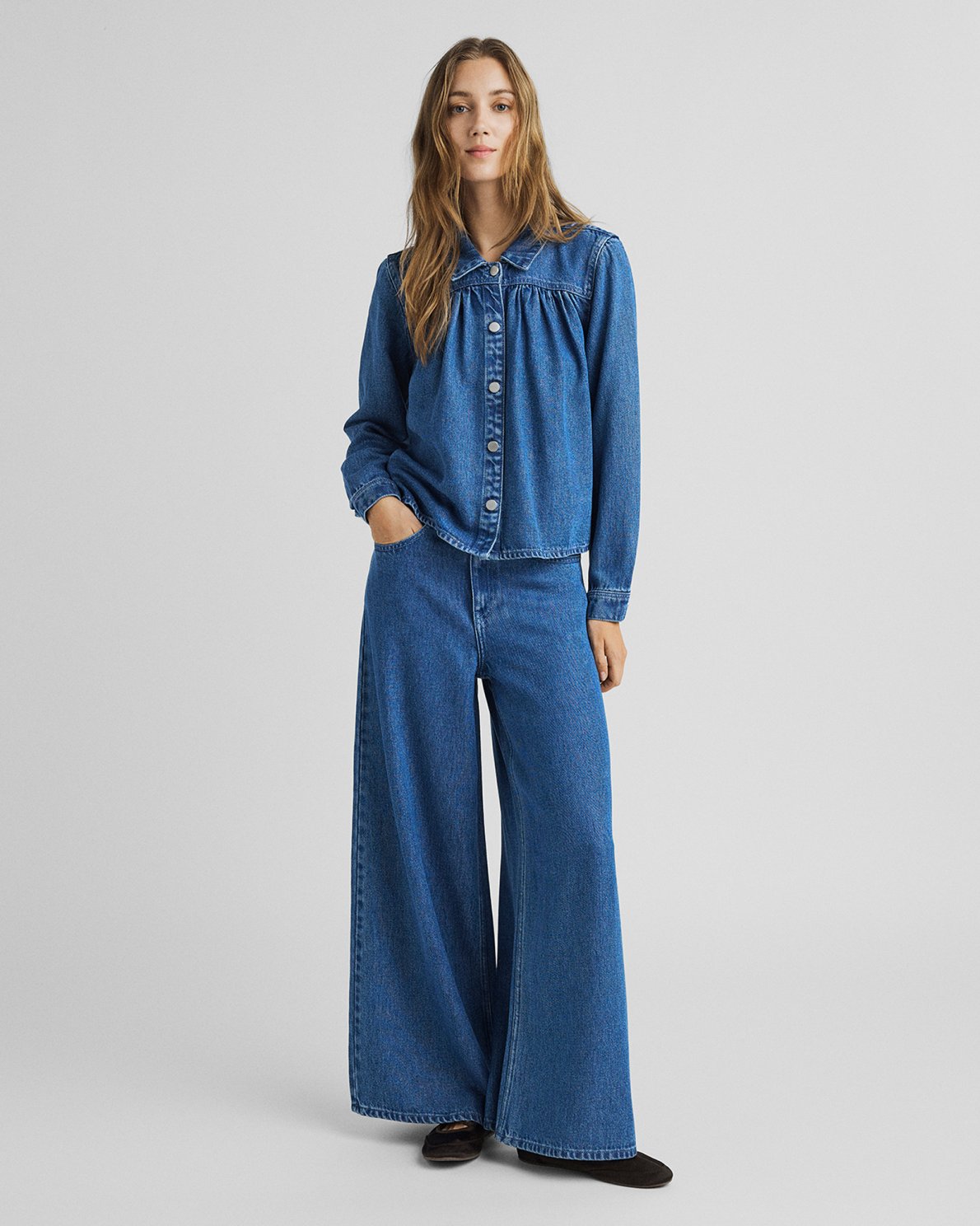 A woman modeling a blue denim set consisting of a cropped button-front jacket and matching wide-leg pants, photographed against a plain light gray studio background.