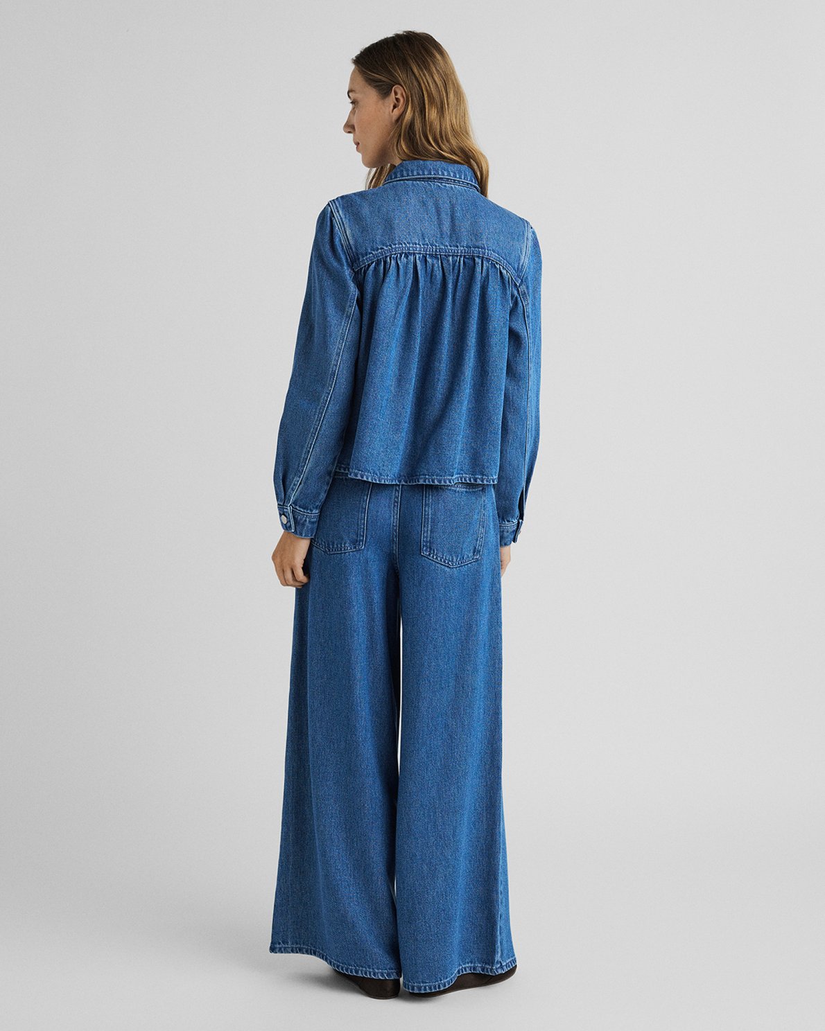 A model shown from the back wearing a blue denim jumpsuit with a cropped, gathered long-sleeve top and wide-leg pants, set against a plain light gray studio background.