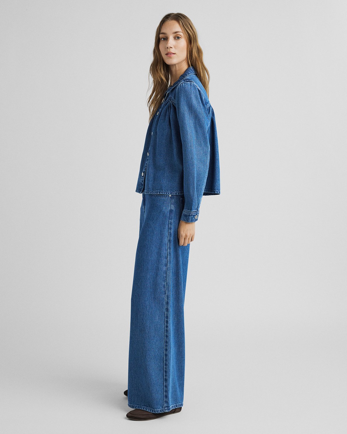 A woman modeling a blue denim set consisting of a cropped button-front jacket and matching wide-leg pants, photographed against a plain light gray studio background.