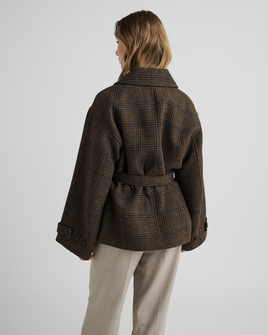A woman shown from the back wearing a brown plaid belted jacket and light beige trousers, photographed against a plain light gray studio background.