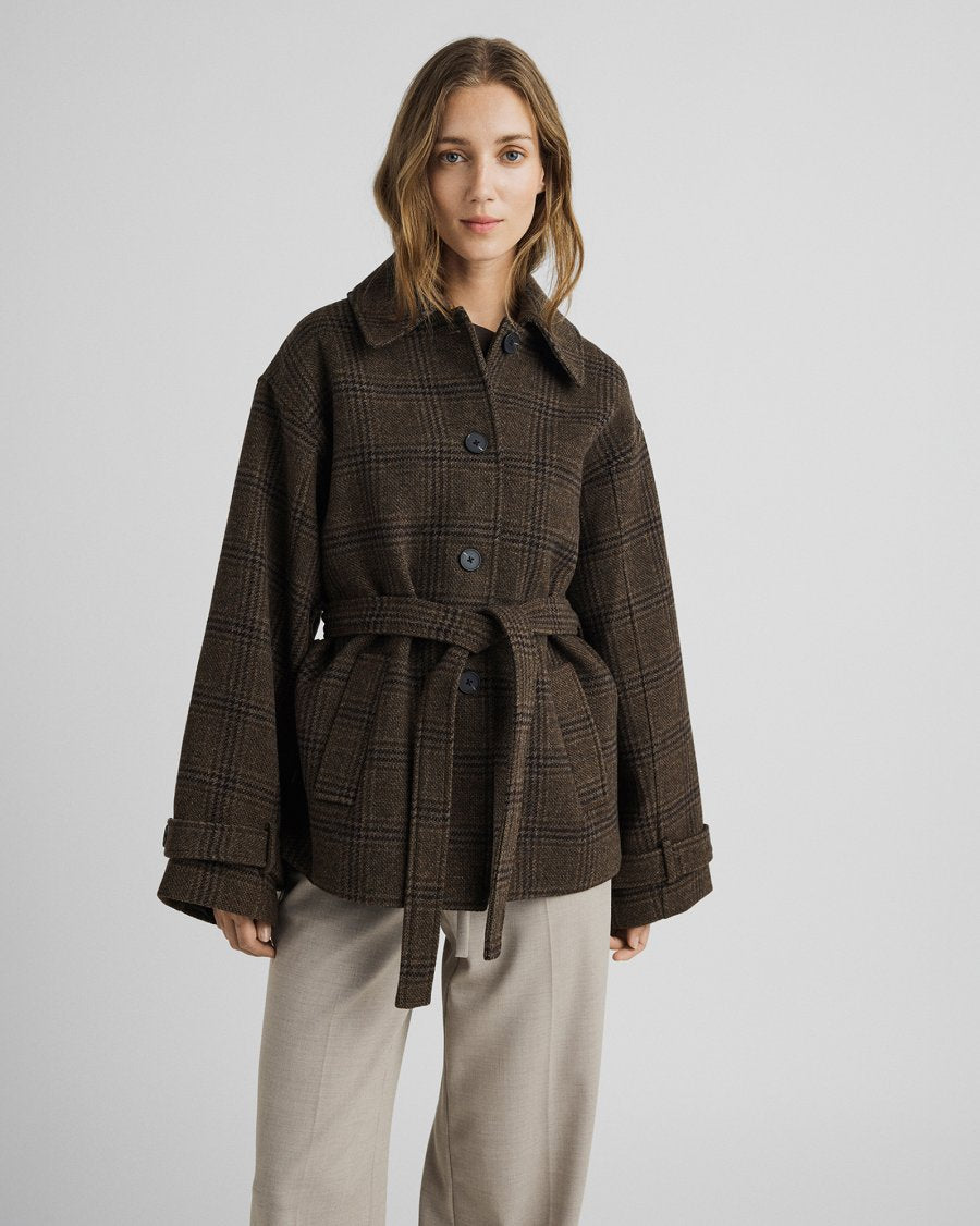 A woman modeling a dark brown plaid belted coat with wide sleeves, paired with light beige trousers, against a neutral gray studio background.