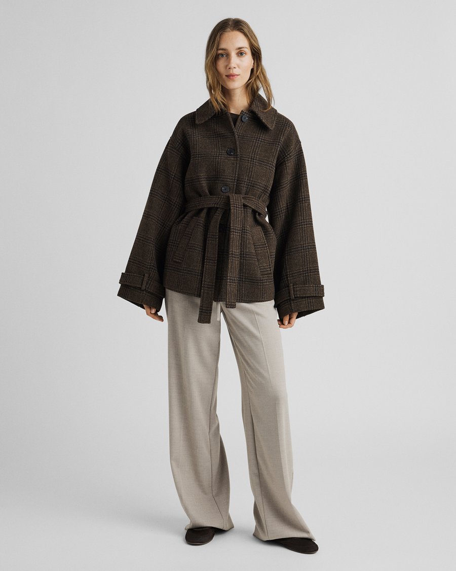 A woman stands against a light gray studio background wearing a dark brown belted plaid coat with wide sleeves, light beige wide-leg trousers, and dark shoes.