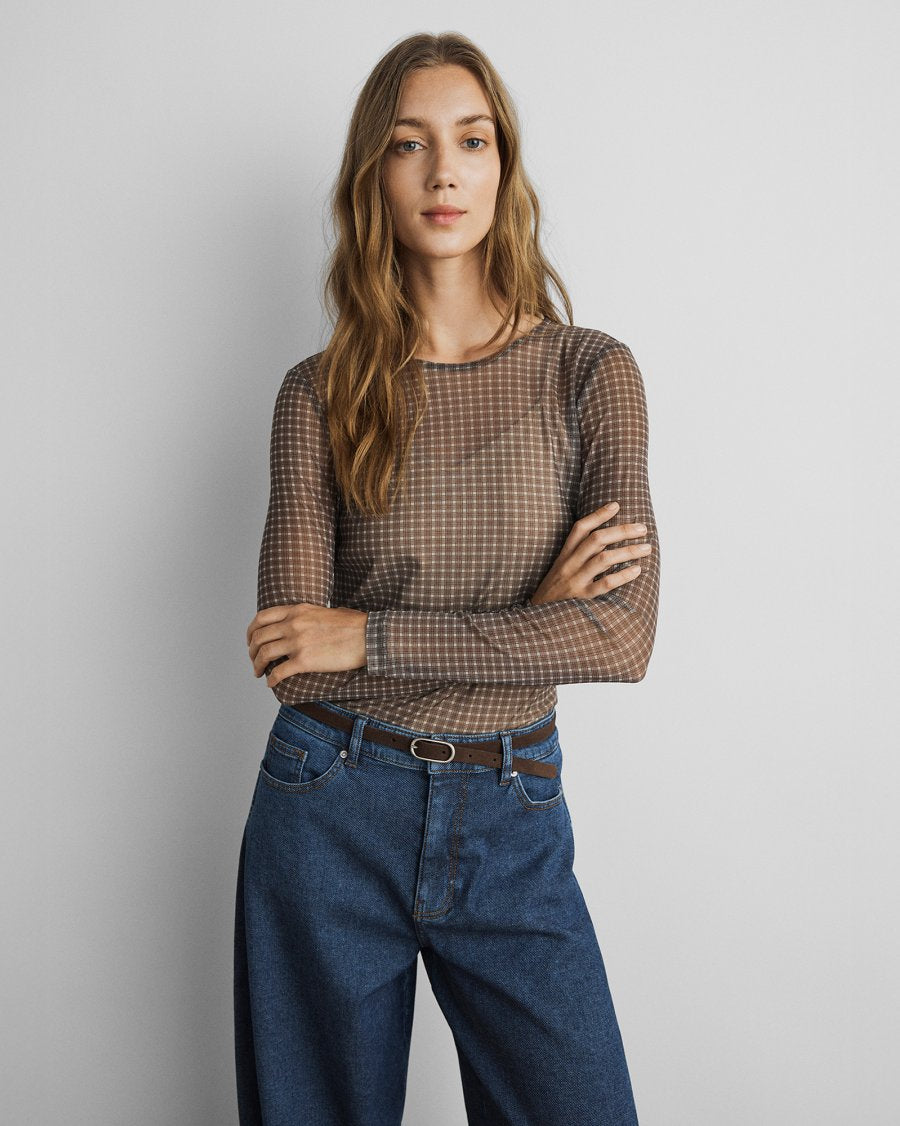 A woman modeling a brown and beige checkered sheer long-sleeve top paired with high-waisted blue jeans and a brown belt, standing against a light gray studio background.