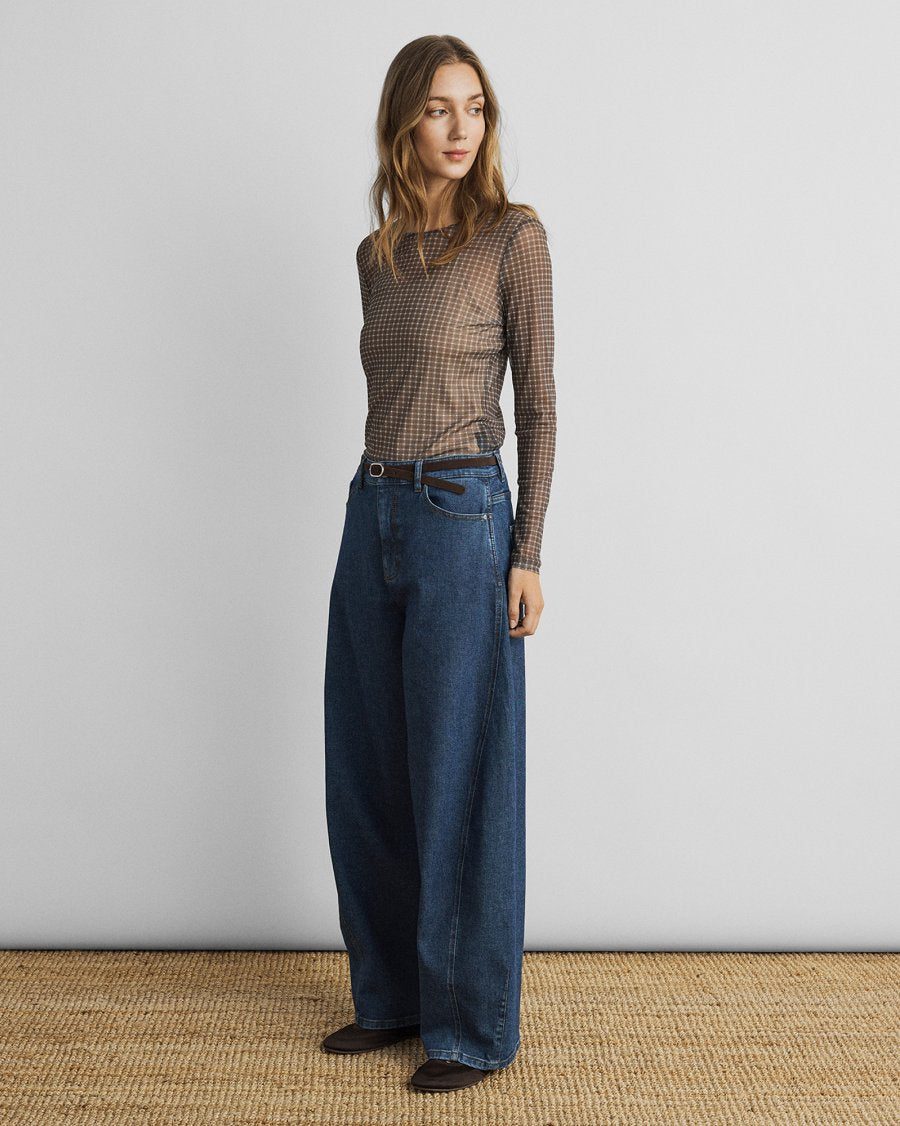 A woman stands in a studio wearing a sheer brown-and-black check long-sleeve top and wide-leg dark blue jeans, with a thin belt and dark shoes, against a light gray wall and woven beige floor.