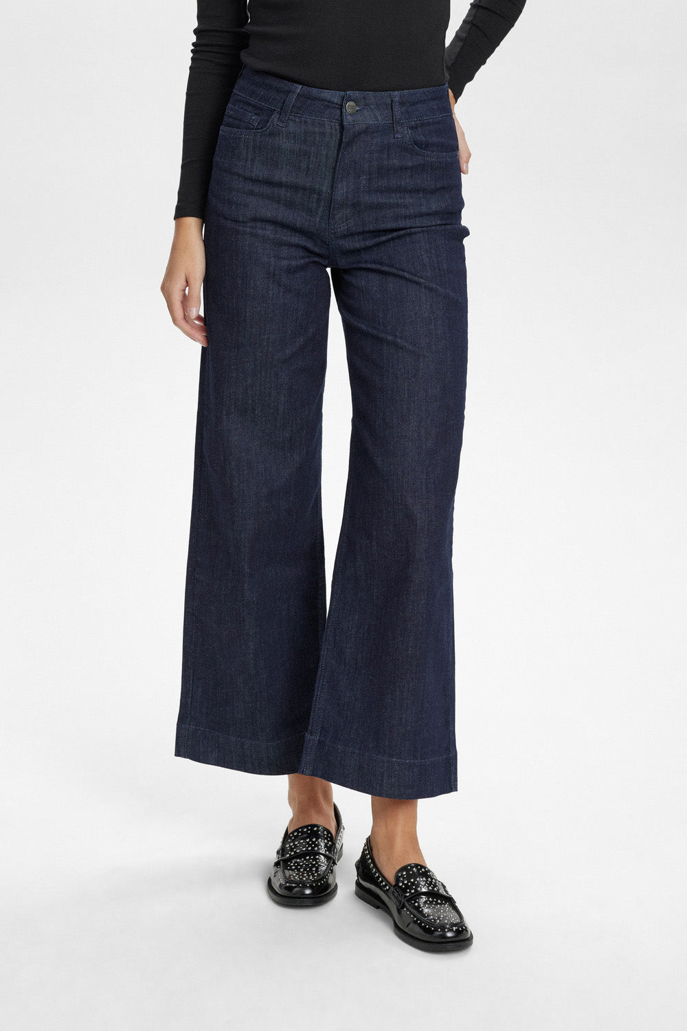 A model wearing dark blue wide-leg cropped jeans, a black long-sleeve top, and black patent leather loafers, photographed against a clean white studio background.