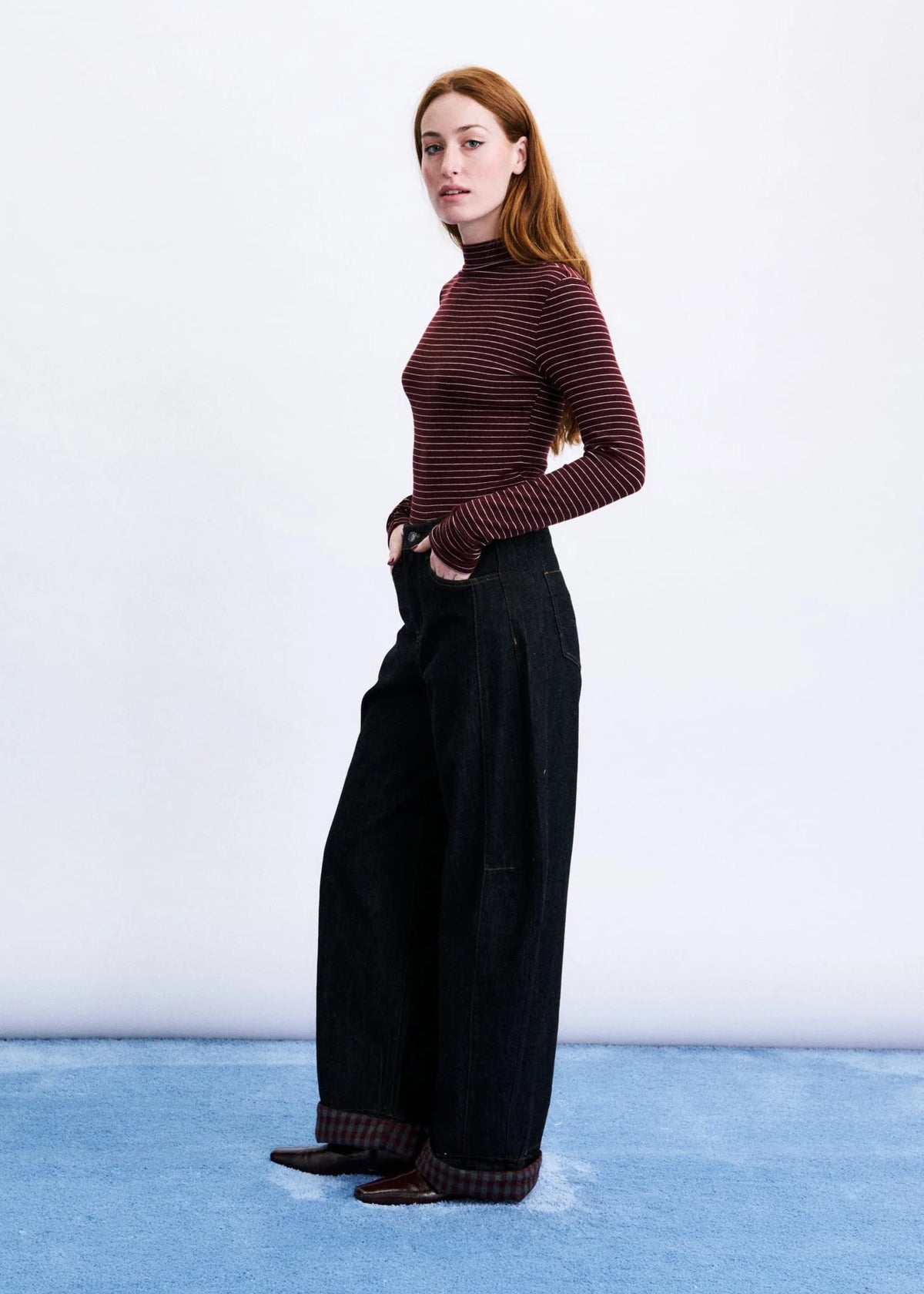 A woman stands in a studio wearing a fitted long-sleeve turtleneck top with thin dark red and black horizontal stripes, paired with very wide-leg black trousers and dark pointed shoes.