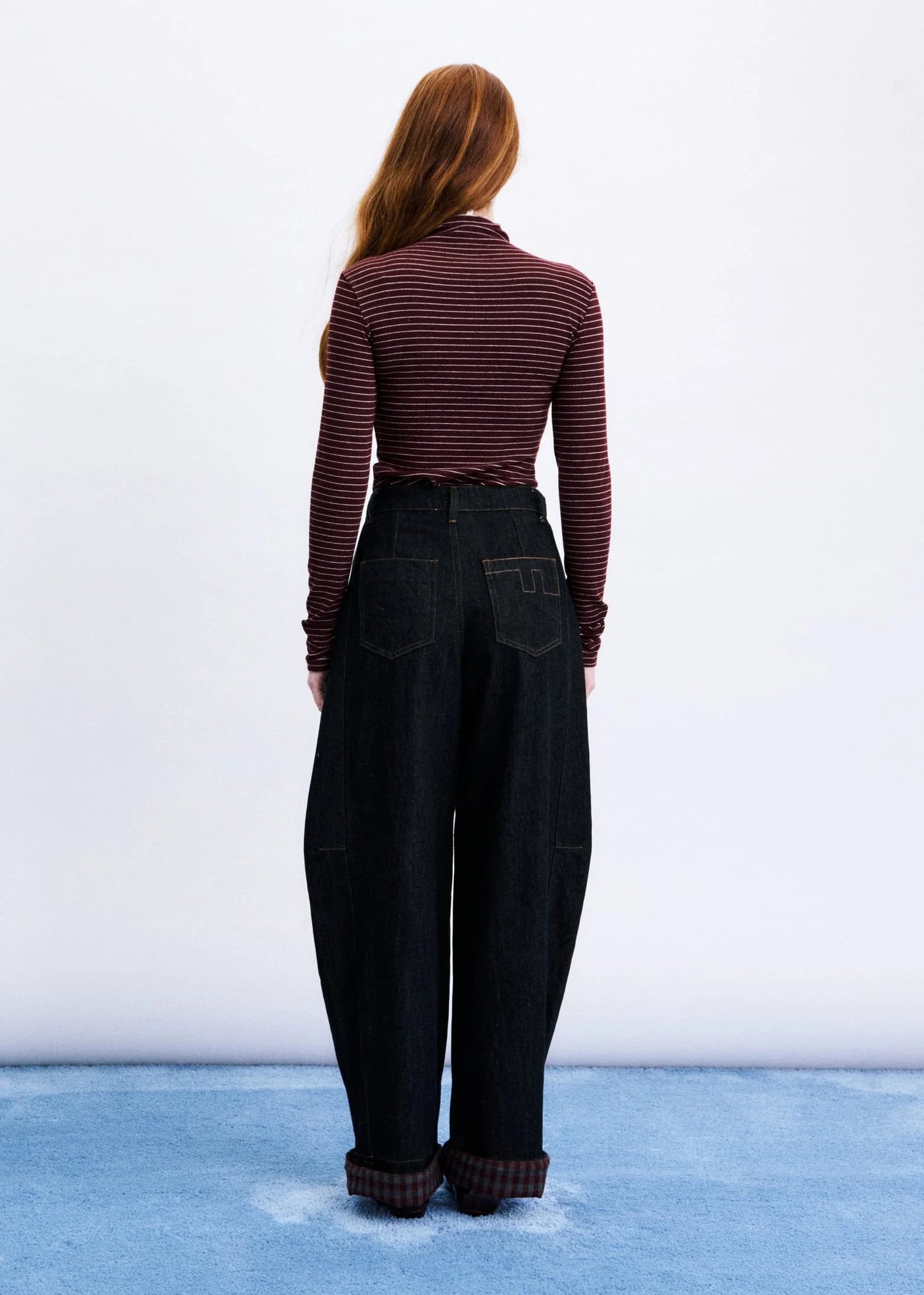 A woman viewed from behind wearing a dark burgundy and black striped long-sleeve mock neck top and dark wide-leg jeans, standing against a white wall with a blue floor.