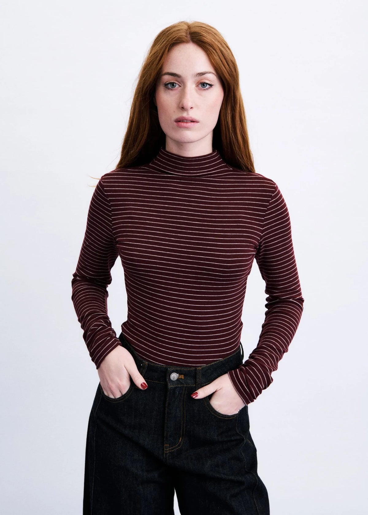 A woman with red hair wearing a dark burgundy and brown striped mock neck long-sleeve top and dark jeans, standing against a light gray background.