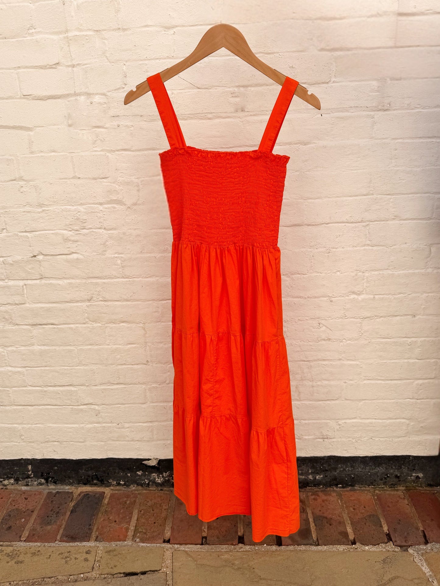 A bright orange tiered midi dress with a smocked bodice and wide shoulder straps, hanging on a wooden hanger against a white painted brick wall.