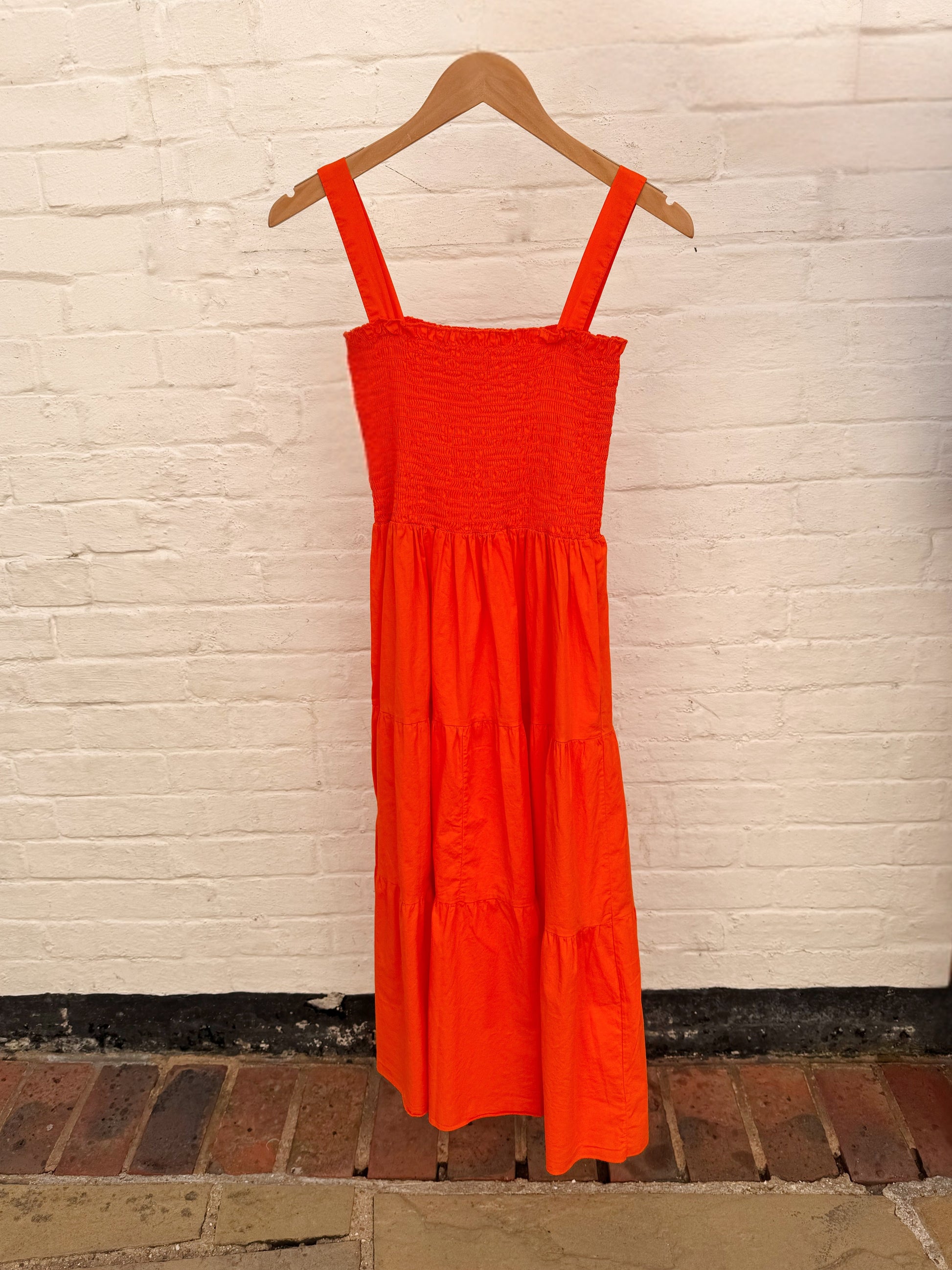 A bright orange tiered midi dress with a smocked bodice and wide shoulder straps, hanging on a wooden hanger against a white painted brick wall.