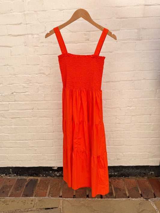 A bright orange tiered midi dress with a smocked bodice and wide shoulder straps, hanging on a wooden hanger against a white painted brick wall.