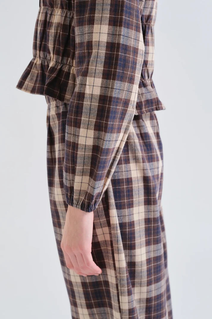 A close-up side view of a person wearing a plaid long-sleeve dress or jumpsuit in brown, beige, and navy tones, with a ruffled sleeve and gathered waist detail, against a plain light gray background.