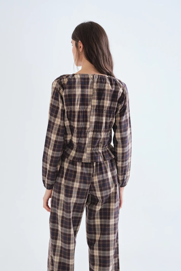 A woman viewed from behind wearing a long-sleeve plaid jumpsuit in dark brown, beige, and black tones, set against a plain light gray studio background.