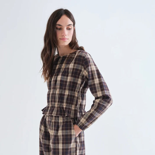 A woman modeling a matching plaid long-sleeve top and wide-leg pants set in a dark brown, beige, and black tartan pattern, set against a plain light gray studio background.