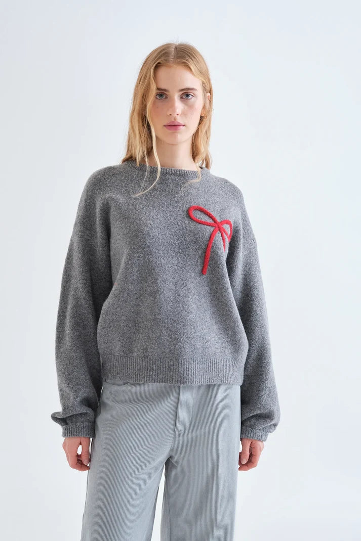 A woman modeling a heather gray knit sweater with a red bow detail on the chest, paired with light gray trousers, against a plain white studio background.
