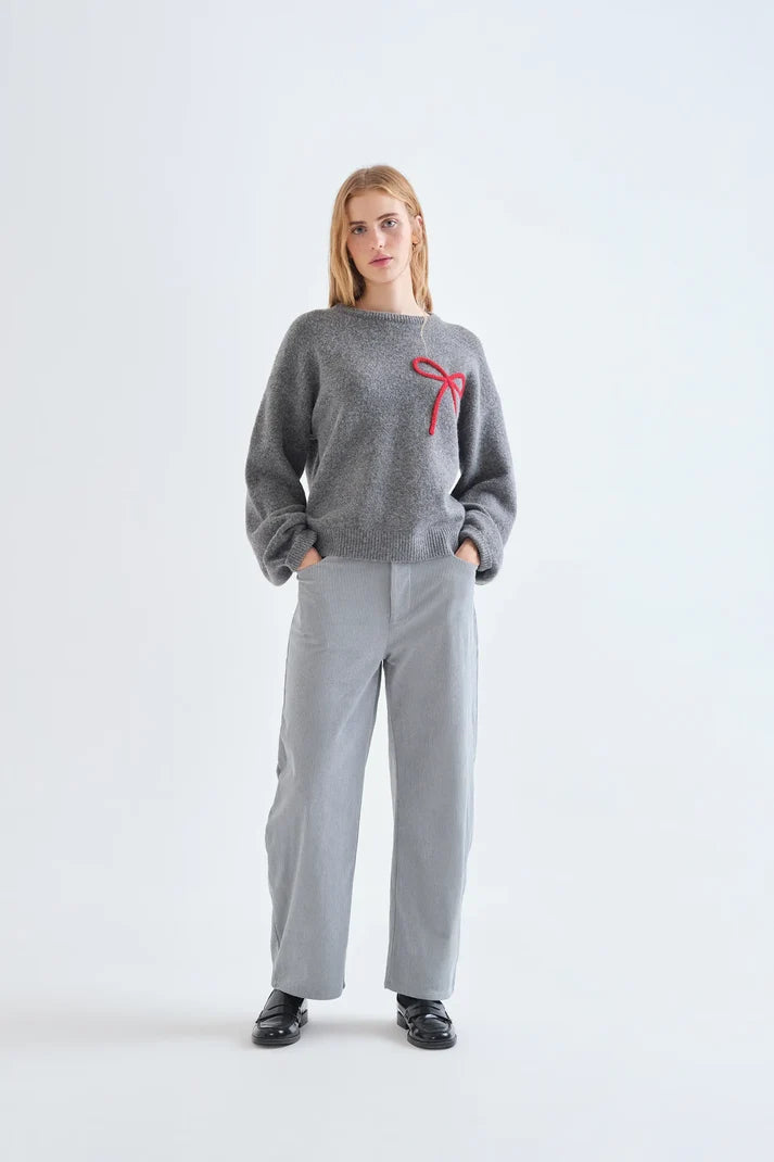 A woman stands against a light gray studio background wearing a fuzzy heather gray crewneck sweater with a red bow detail on the chest, paired with light gray wide-leg trousers and black shoes.
