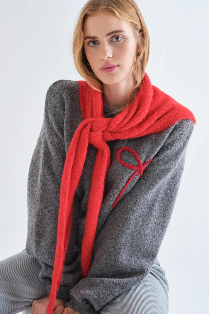 A woman wearing a chunky gray knit sweater and a bright red knit scarf tied around her neck, photographed against a clean white studio background.