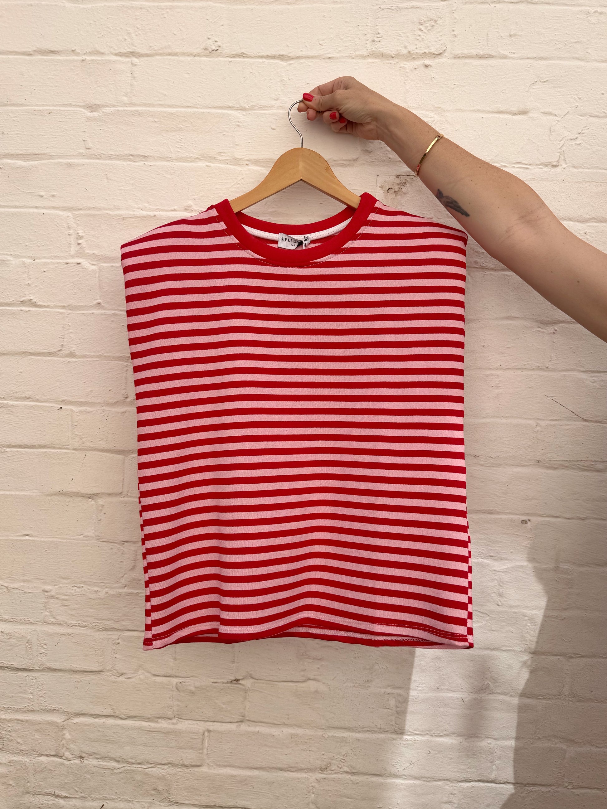 A red and pink horizontally striped sleeveless top hanging on a wooden hanger against a white painted brick wall, with a person's arm holding the hanger hook.