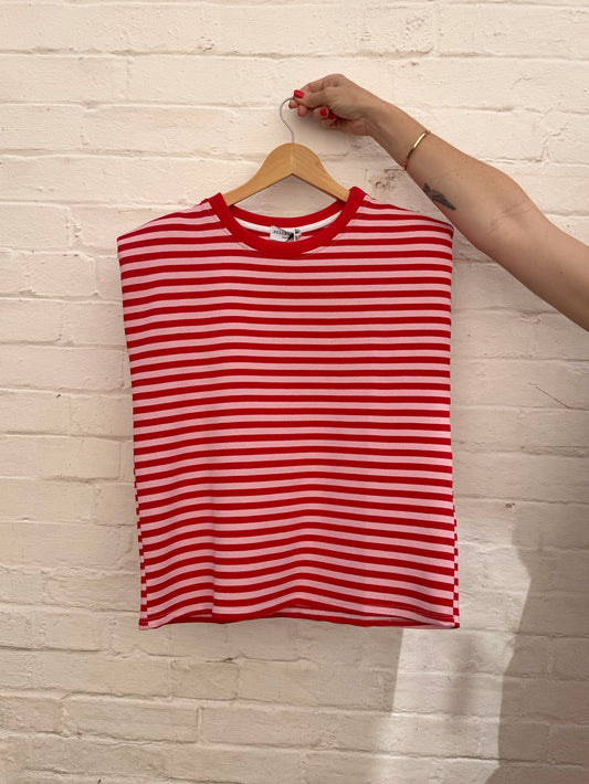 A red and pink horizontally striped sleeveless top hanging on a wooden hanger against a white painted brick wall, with a person's arm holding the hanger hook.