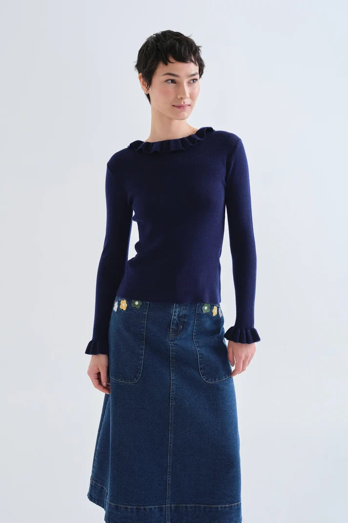 A model wearing a navy blue long-sleeve knit top with a ruffled neckline and cuffs, paired with a dark blue denim skirt, against a light gray studio background.