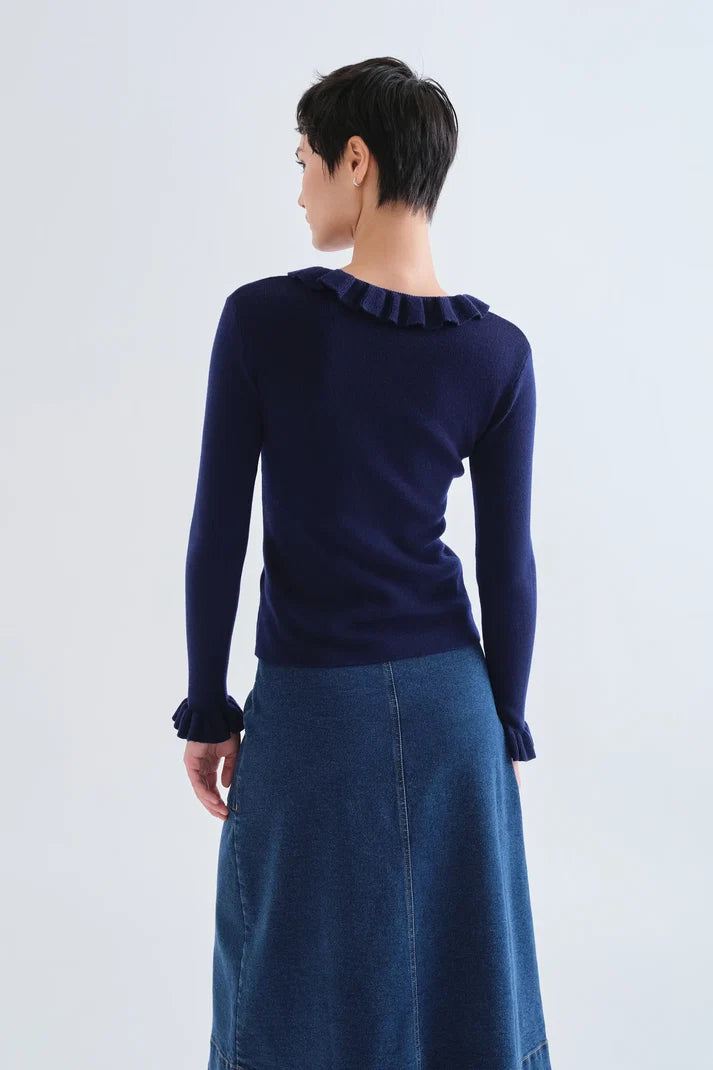 A model shown from the back wearing a navy blue long-sleeve knit top with a ruffled collar and cuffs, paired with a blue denim skirt, against a light gray studio background.
