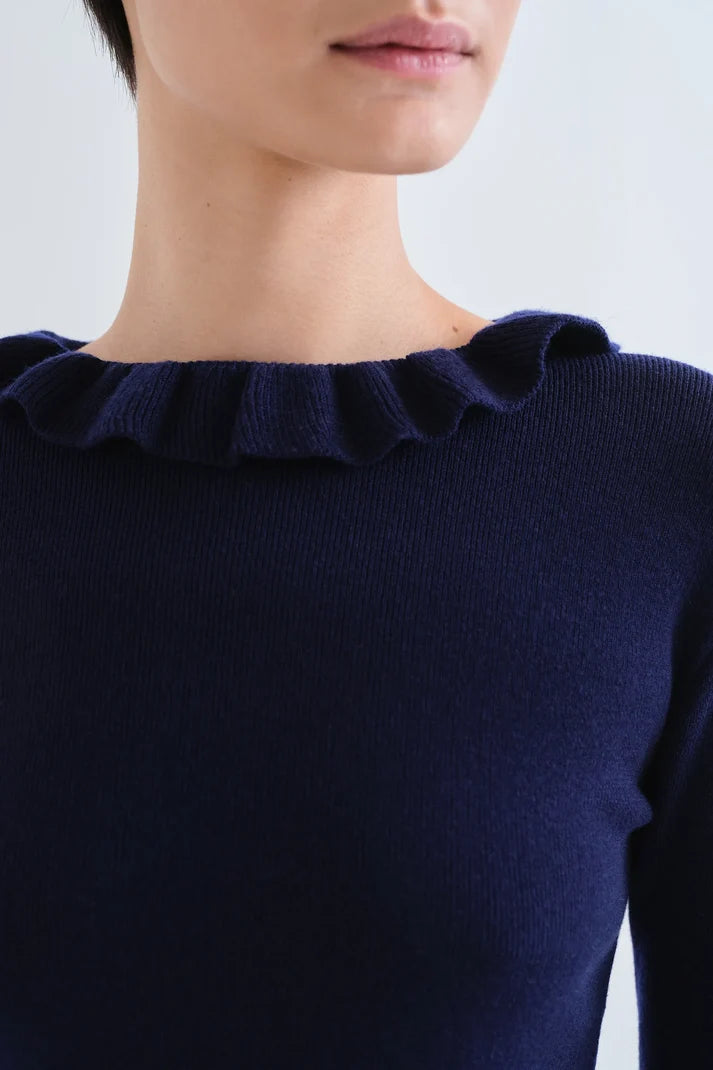 Close-up of a model wearing a dark navy blue ribbed knit top with a ruffled boat neckline against a light gray background.