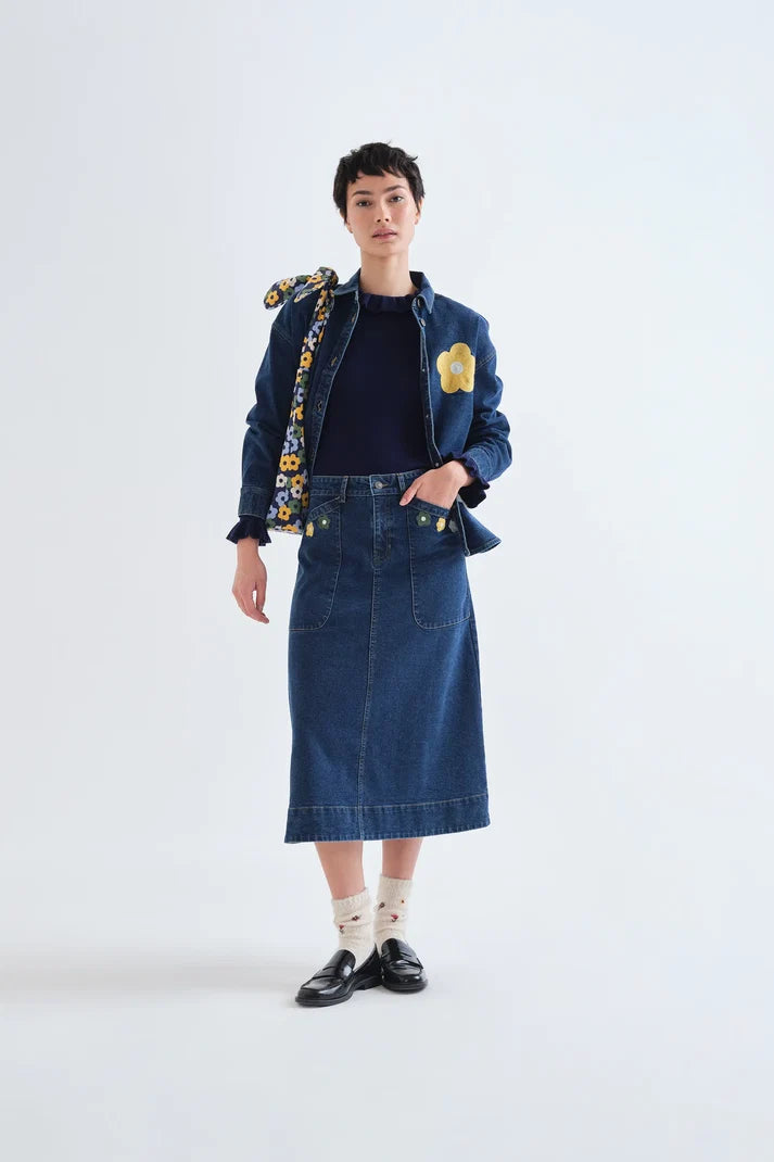 A model stands against a light gray studio background wearing a dark blue denim jacket with yellow floral embroidery and a matching blue denim midi skirt. The outfit is styled with a dark navy top, white socks, and black loafers.