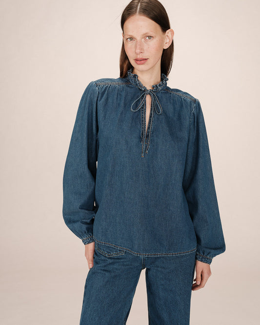 Woman wearing a blue denim blouse with long sleeves and a tie neckline on a beige background