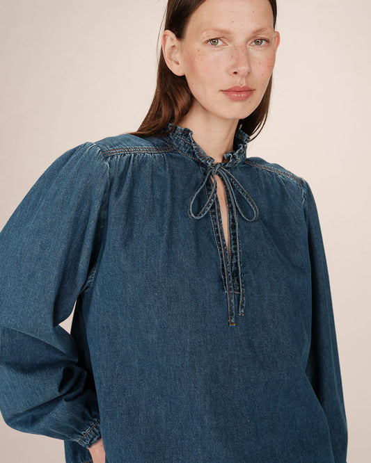 Woman wearing a blue denim blouse with a tie neckline against a beige background