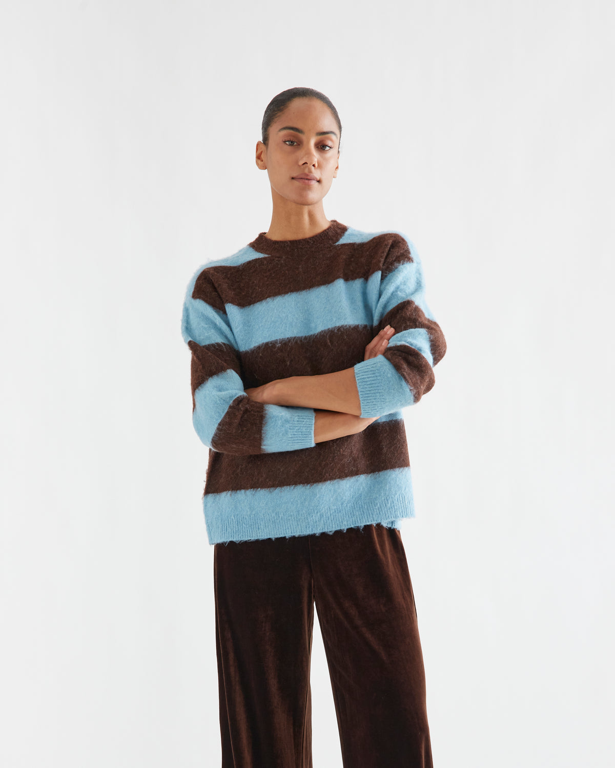 A model wearing a fuzzy, long-sleeve knit sweater with horizontal brown and light blue stripes, paired with dark brown velvet trousers, against a plain white studio background.