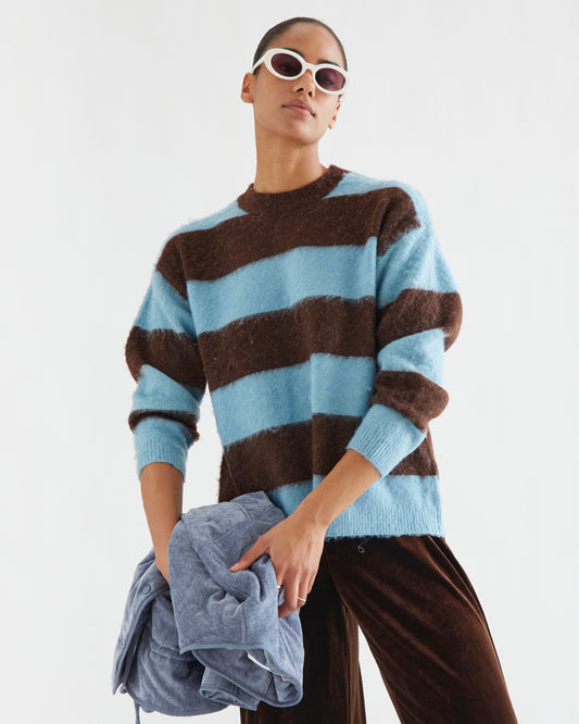 A model wearing a fuzzy, oversized crewneck sweater with horizontal light blue and dark brown stripes, styled with brown velvet trousers and white-framed sunglasses, holding a folded blue garment against a plain white studio background.