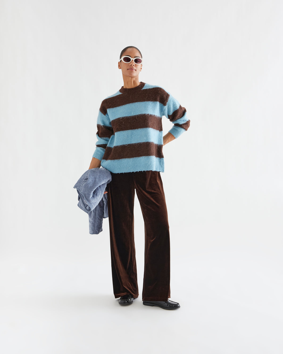 A model stands against a plain white studio background wearing a fuzzy knit sweater with horizontal brown and light blue stripes, dark brown wide-leg velvet pants, black shoes, and white sunglasses, while holding a light blue jacket.
