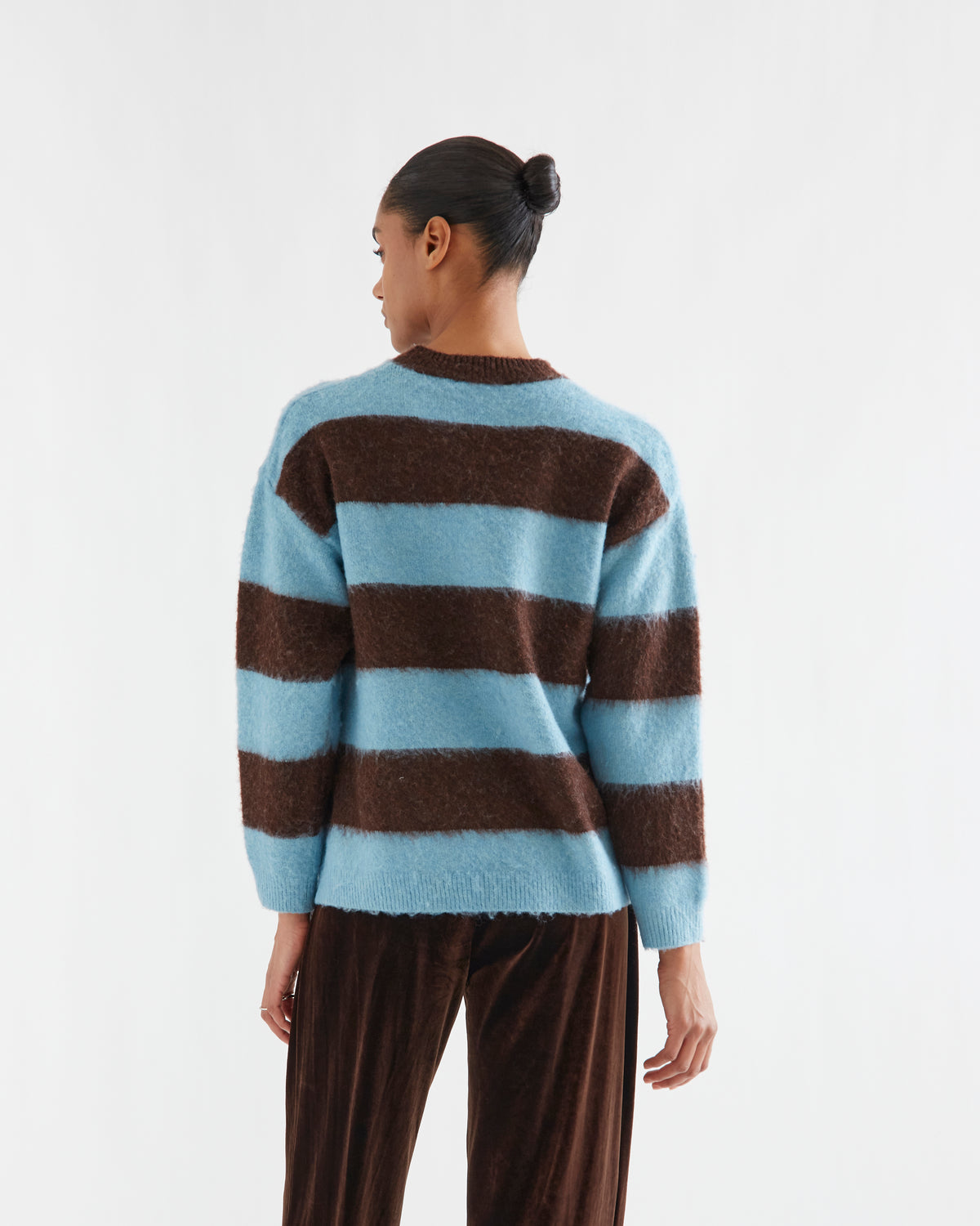 A model shown from the back wearing a fuzzy knit sweater with horizontal light blue and dark brown stripes, paired with brown pants, against a plain white studio background.