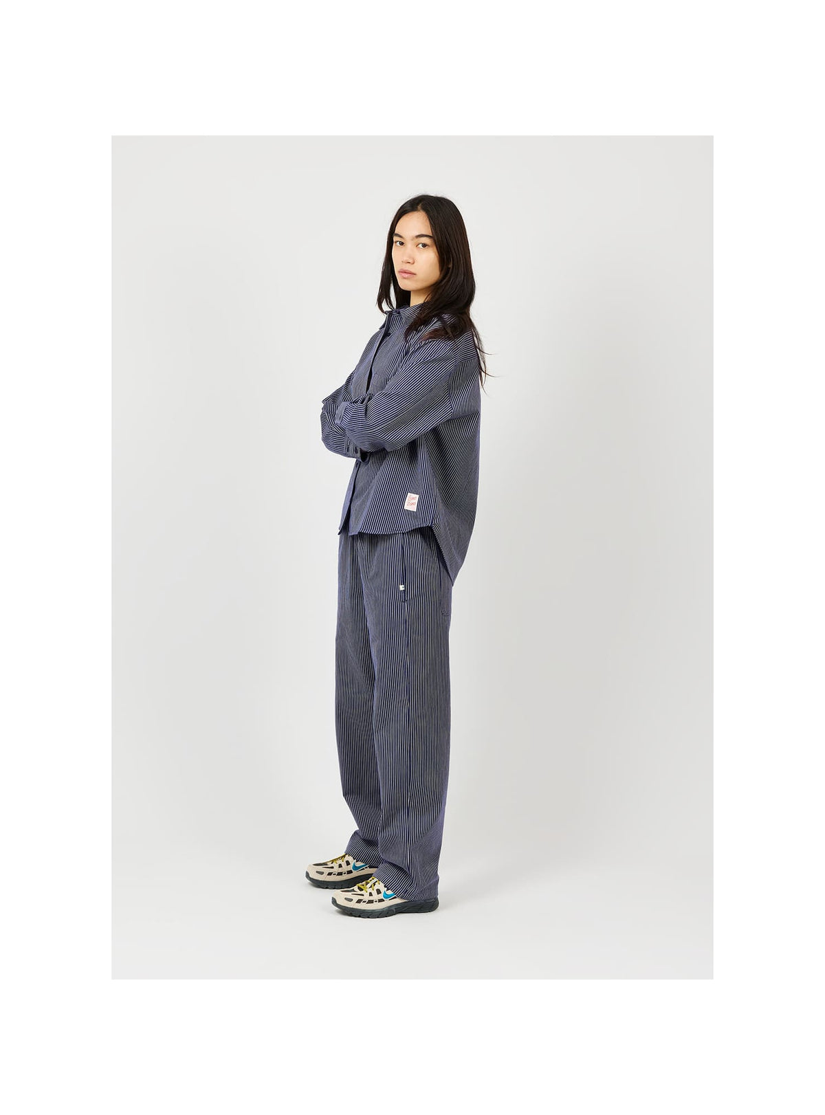 A model stands in a studio wearing a matching dark slate blue lounge set consisting of a long-sleeve button-up shirt and wide-leg trousers, paired with patterned slip-on shoes.