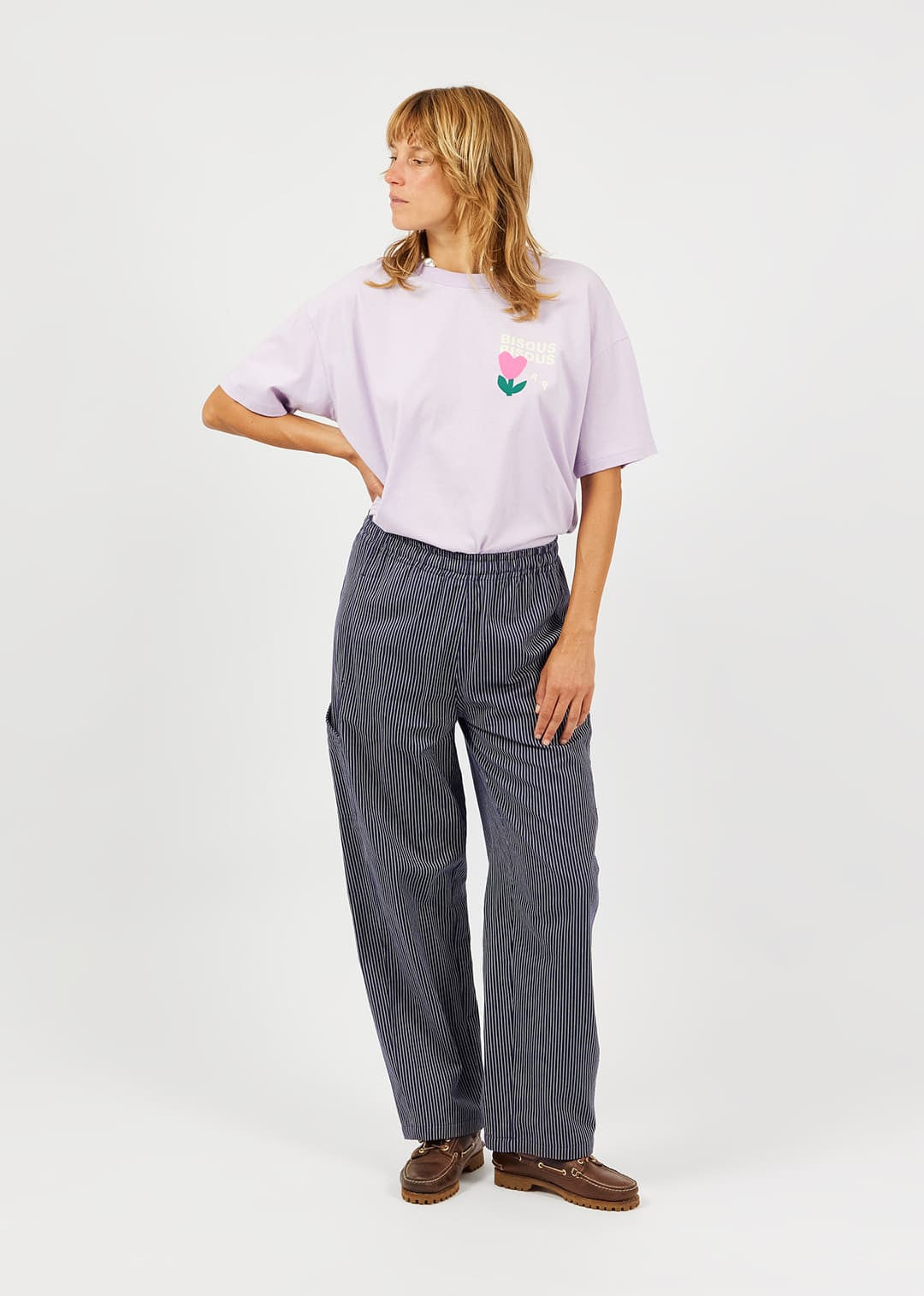 A woman modeling a light purple t-shirt with a small pink and green tulip graphic on the left chest, paired with dark gray wide-leg trousers and brown leather shoes, against a plain white studio background.