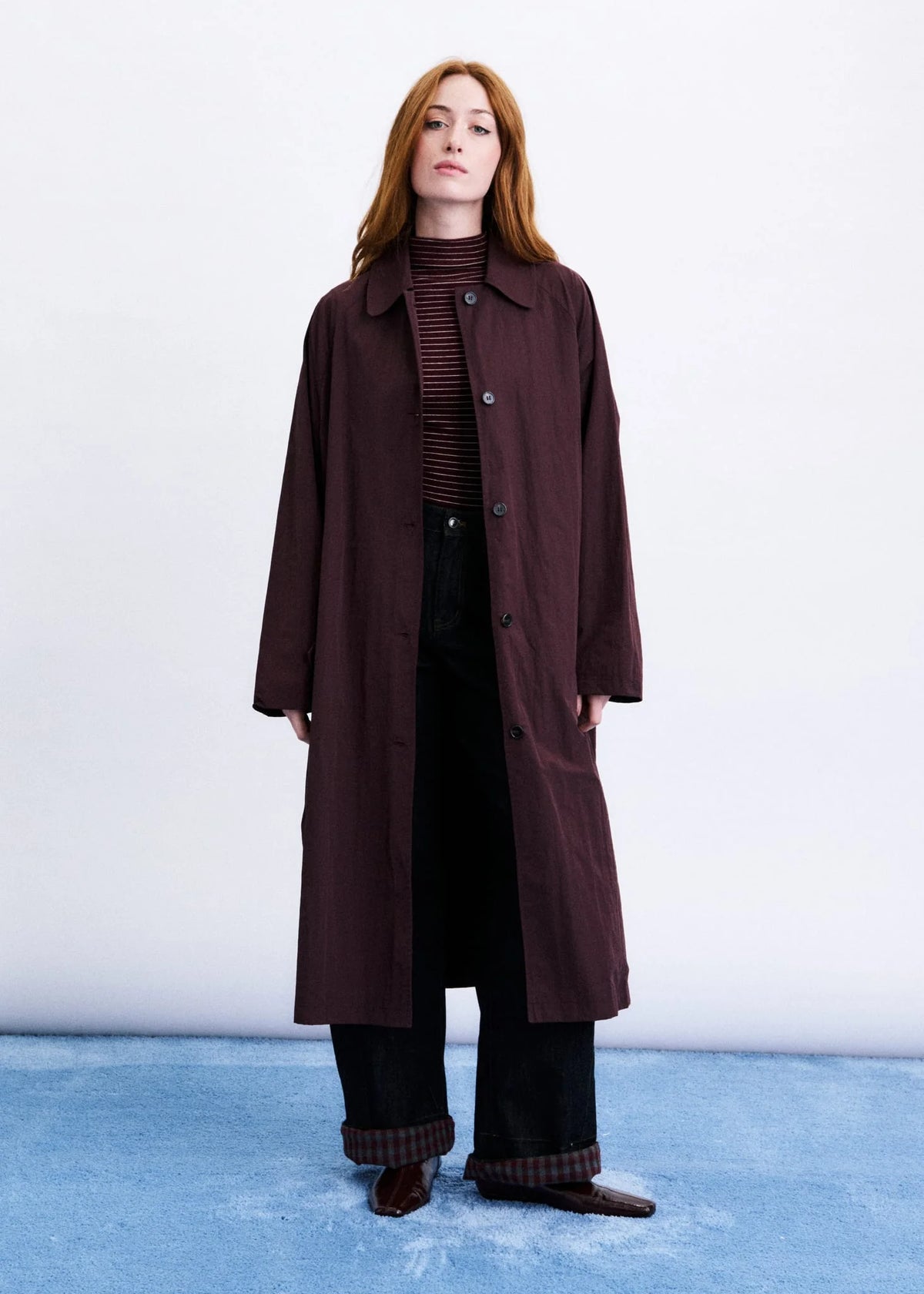 A woman stands against a white studio wall wearing a long dark burgundy coat over a dark striped turtleneck, black wide-leg pants, and brown shoes.