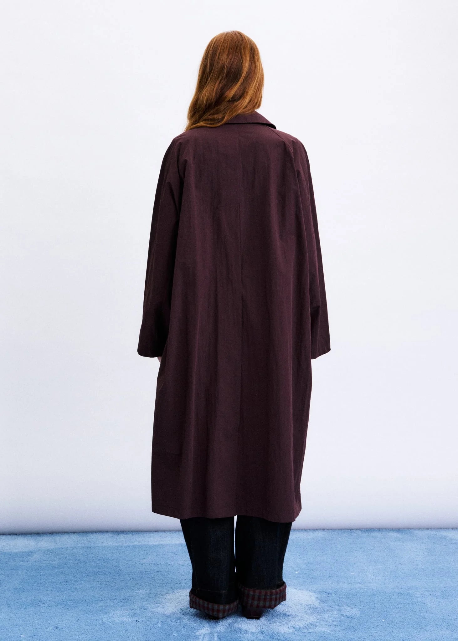 A woman viewed from behind wearing a long, dark plum-brown coat over dark pants, standing against a white studio background with a blue floor.