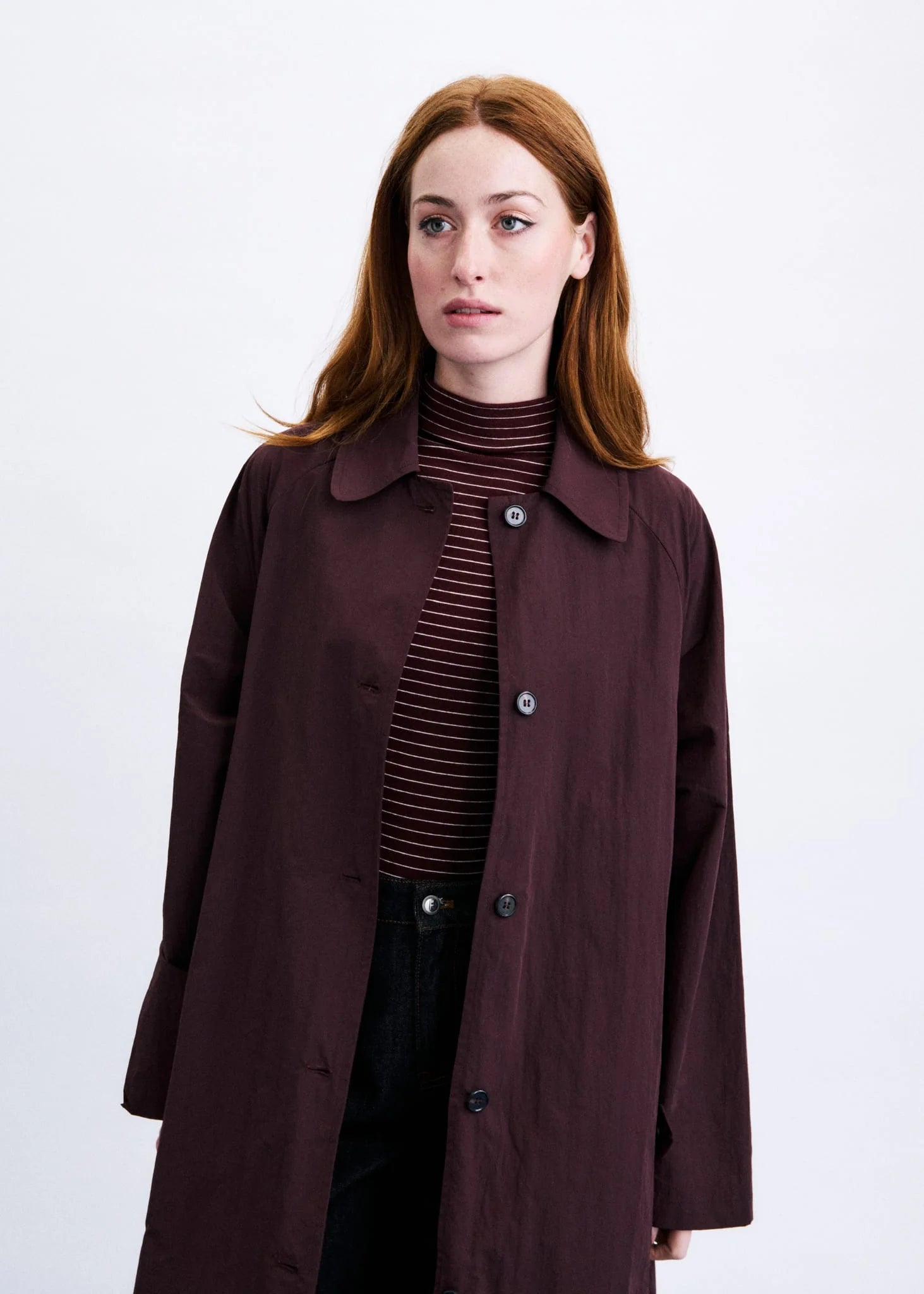 A woman with red hair wearing a dark plum-brown trench coat over a dark striped turtleneck sweater and black pants, photographed against a plain light gray studio background.