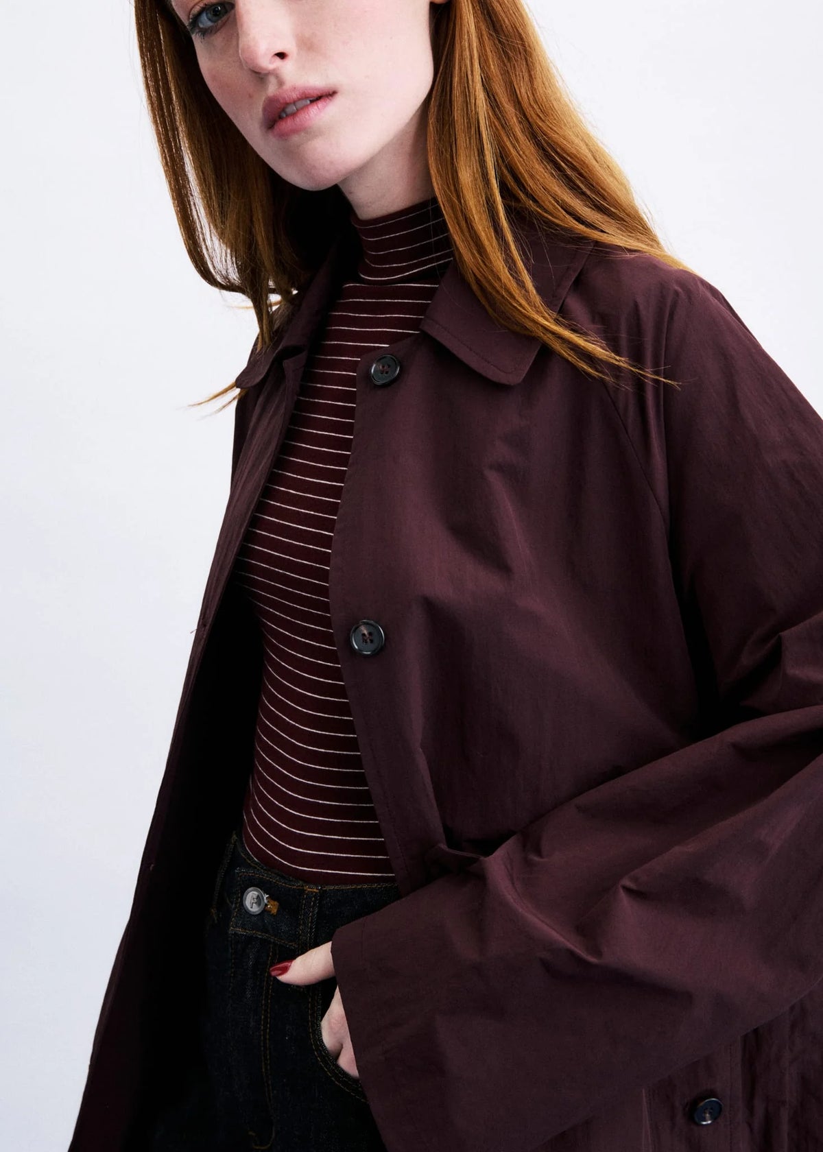 A close-up of a model wearing a dark burgundy oversized button-up coat over a matching burgundy and black striped turtleneck sweater and dark jeans.