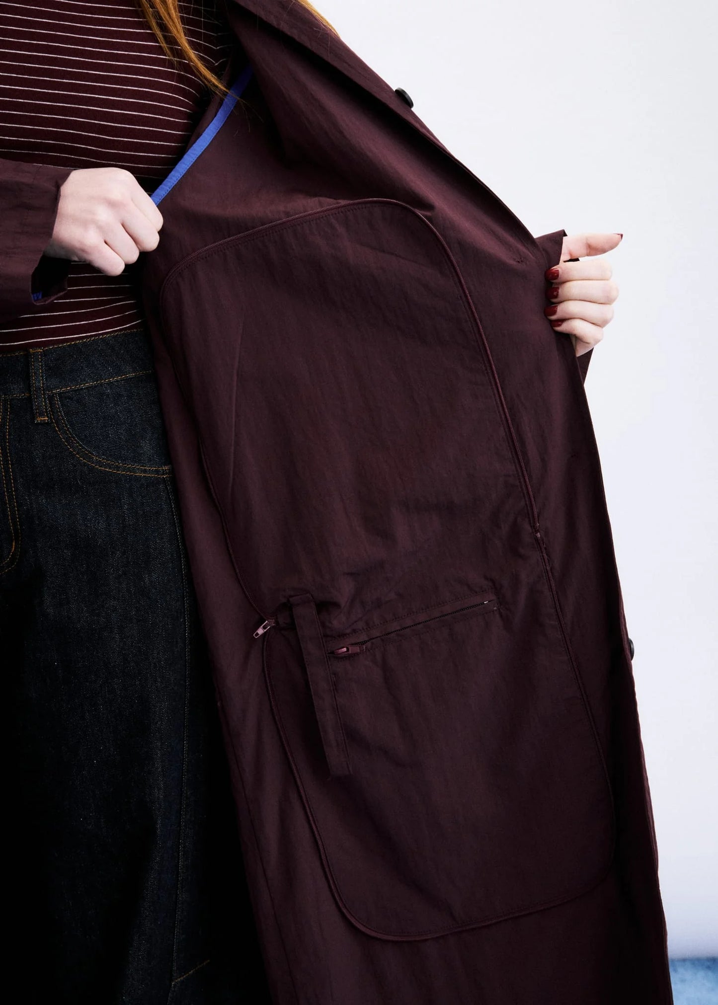 A close-up of a person wearing a dark burgundy trench coat, holding it open to show the interior lining and a large front pocket. The person is also wearing a striped top and dark jeans.