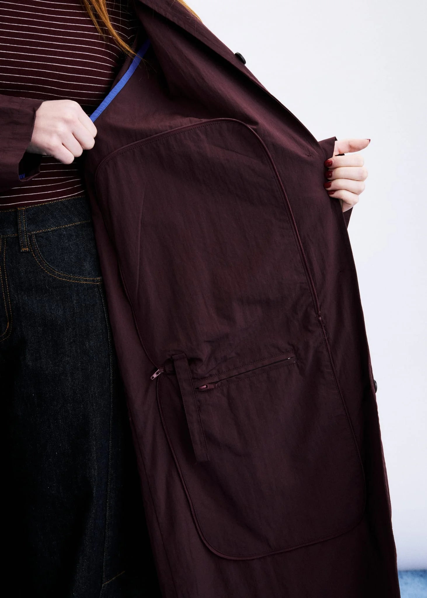 A close-up of a person wearing a dark burgundy trench coat, holding it open to show the interior lining and a large front pocket. The person is also wearing a striped top and dark jeans.