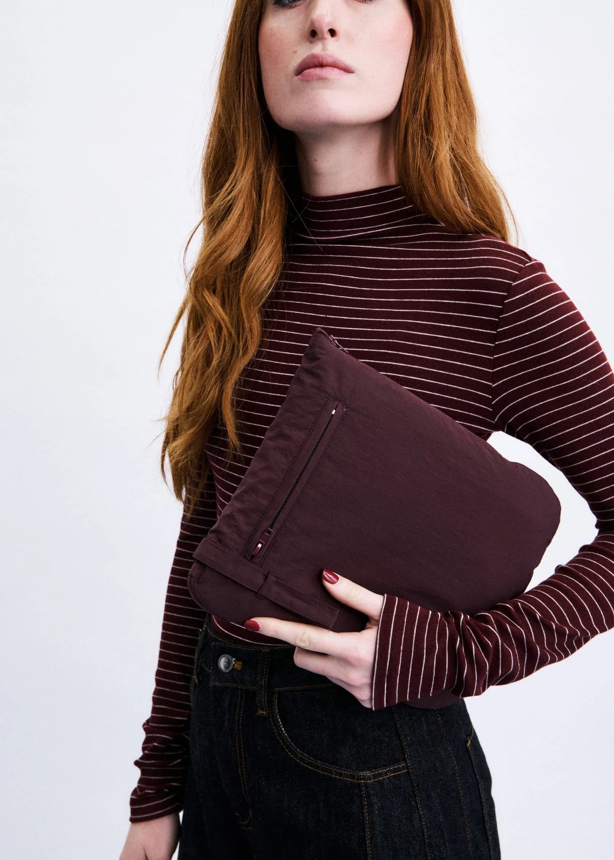 A woman with long red hair holds a dark burgundy fabric pouch or small bag against her chest, wearing a burgundy and white striped mock-neck top and dark jeans, photographed against a light gray studio background.