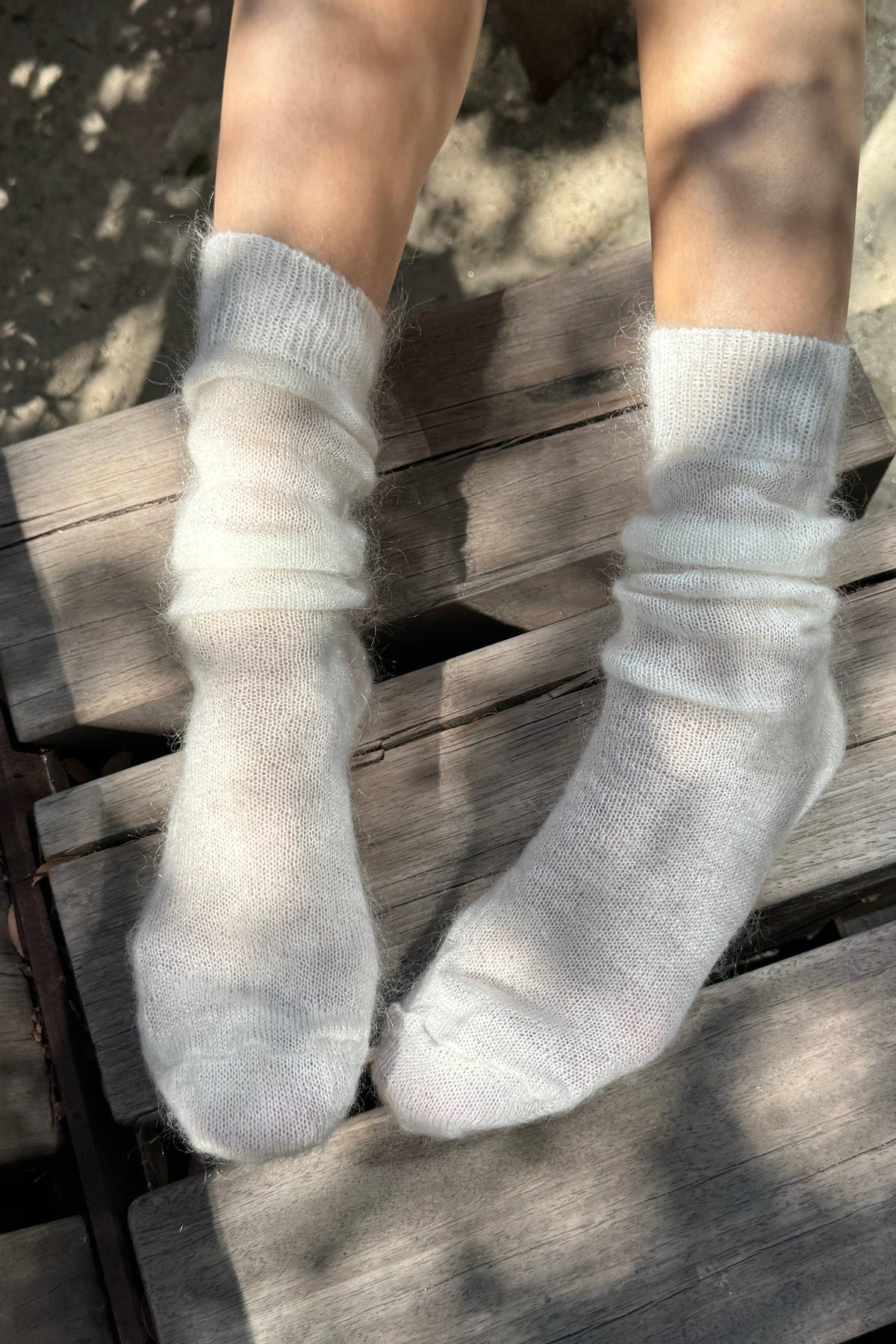 A person wearing fluffy off-white knit socks, sitting on weathered wooden boards outdoors with dappled sunlight and shadows.