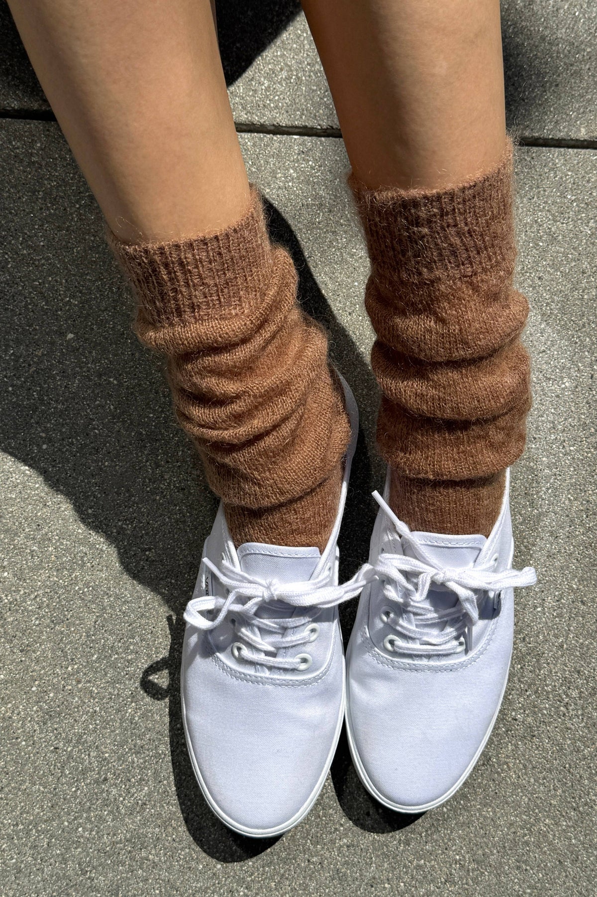 A person wearing brown ribbed knit leg warmers or sock-like boot covers with white low-top sneakers, standing on a gray paved surface.